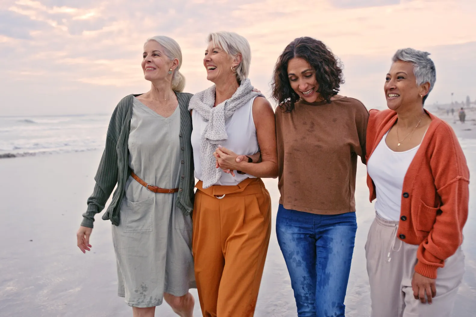 Four women in their 50s and 60s walking together and laughing on a beach at sunset