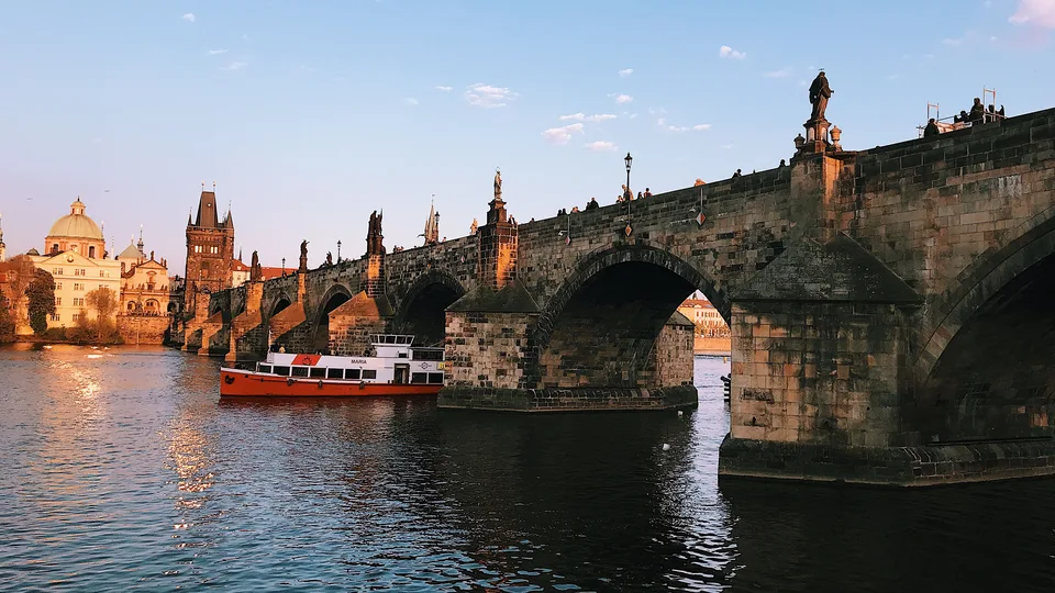 Charles Bridge over the Vltava River at sunset, with Prague Castle on the hill in the distance and a boat passing beneath the bridge